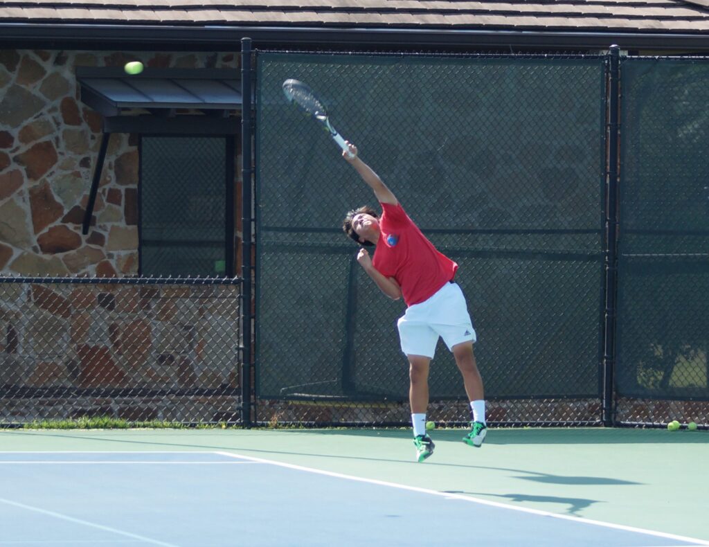 A tennis player in a red shirt serves the ball on an outdoor court beside a stone building.