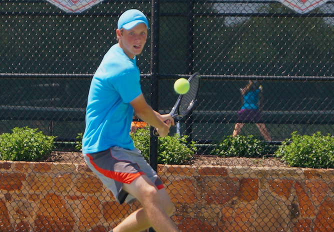 A young man in blue athletic wear hits a tennis ball with a racket on an outdoor court.