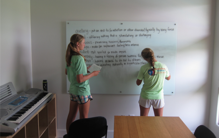 Two girls writing on a whiteboard with vocabulary definitions in a bright classroom with a keyboard nearby.