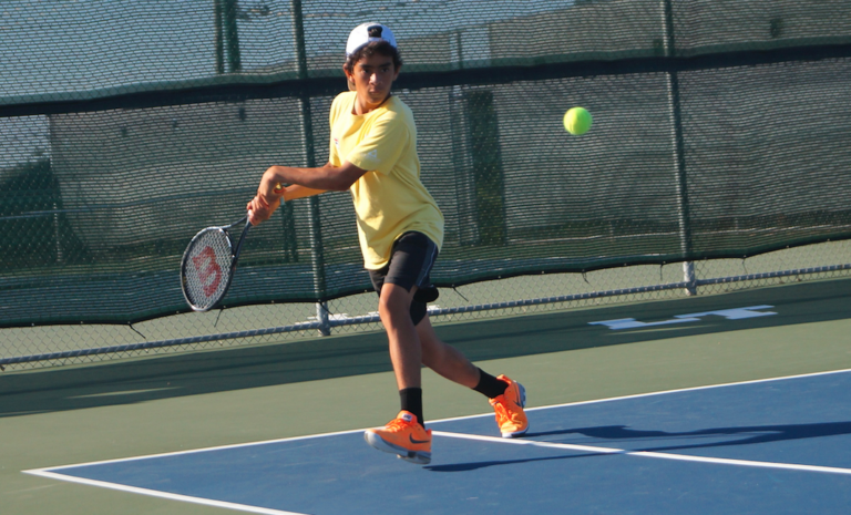 Teen boy in yellow shirt and orange shoes hitting a tennis ball with a racket on an outdoor court.