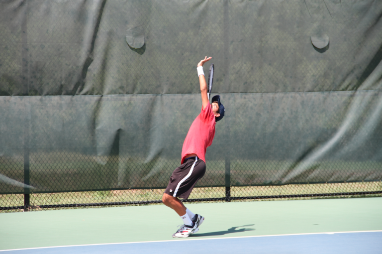A tennis player in a red shirt prepares to serve on an outdoor court with a green fence in the background.