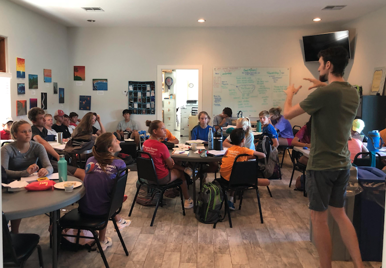 A group of students sit at tables listening to a standing instructor in a classroom with colorful art on the walls.