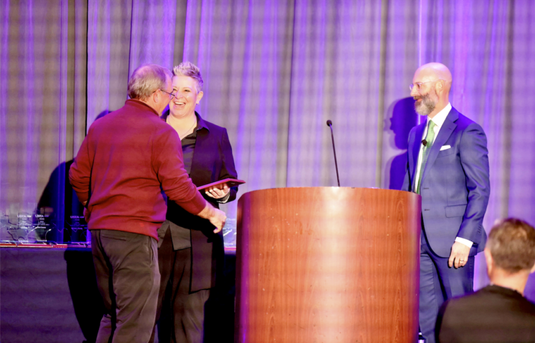 Three people on stage at an award ceremony; one is handing out a plaque while another stands at a podium.