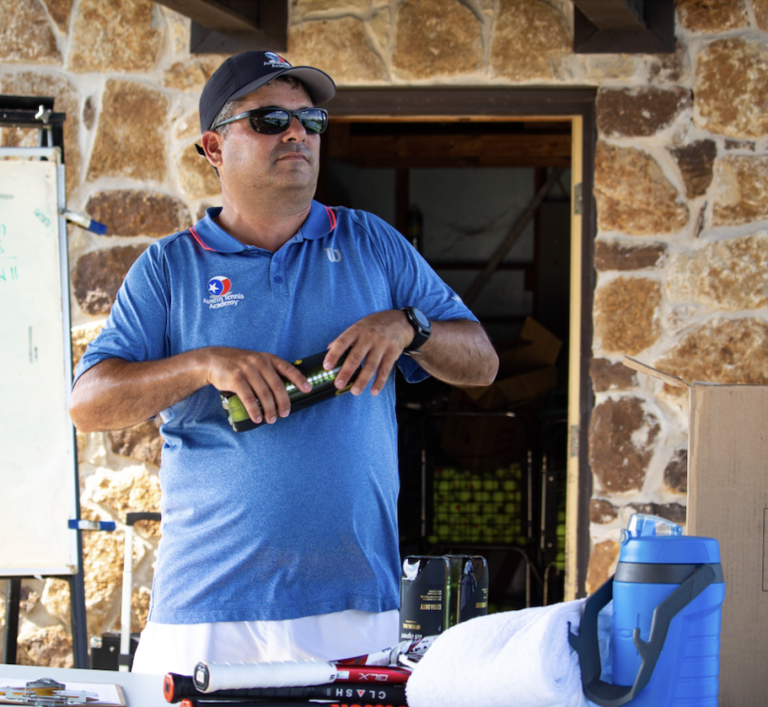 Man in a blue polo and sunglasses stands by a table with sports gear and a water bottle outdoors.