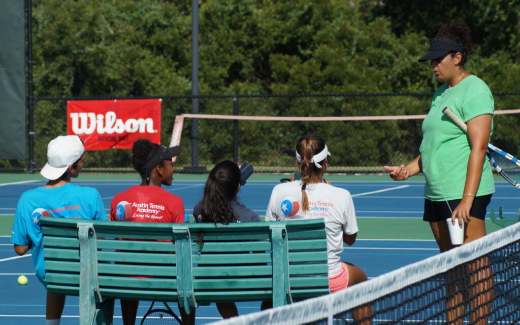 A tennis coach talks to four young players sitting on a bench by a tennis court on a sunny day.