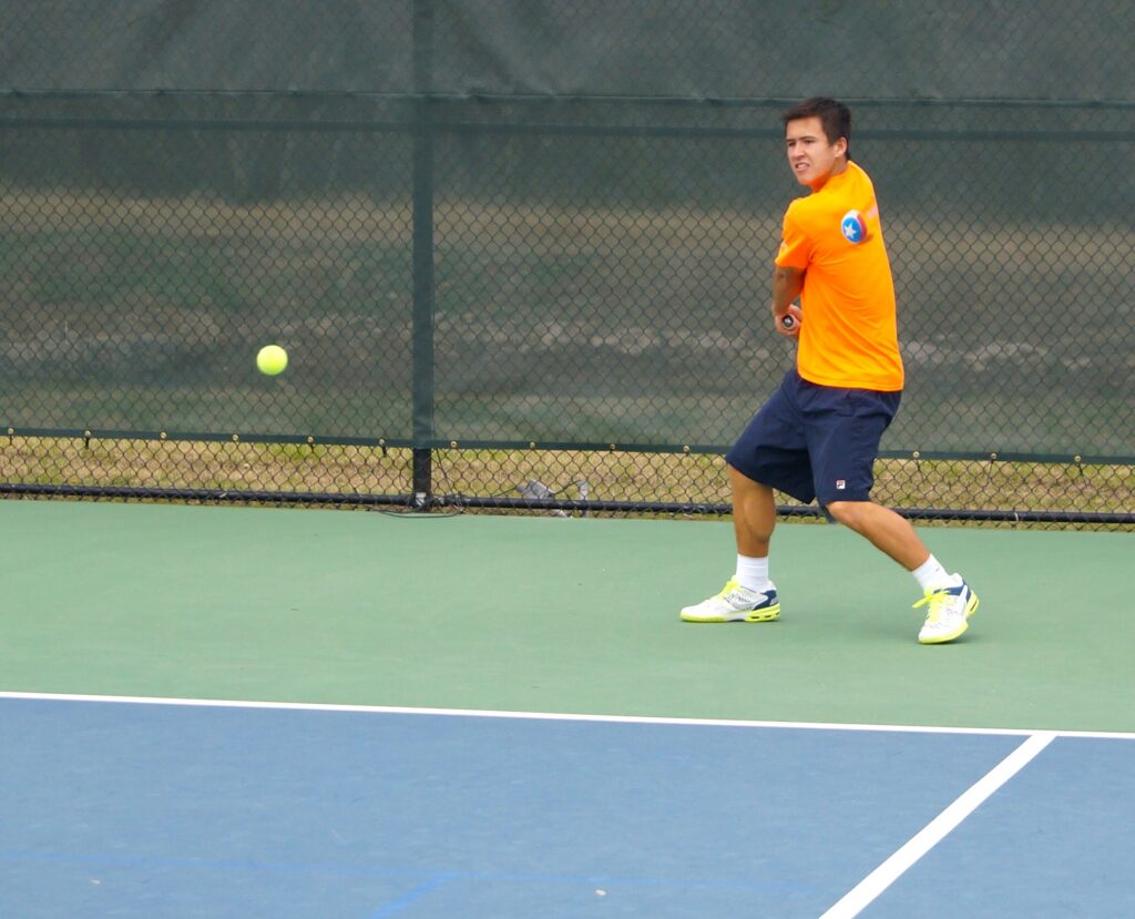 A tennis player in an orange shirt prepares to hit a backhand shot on an outdoor court.