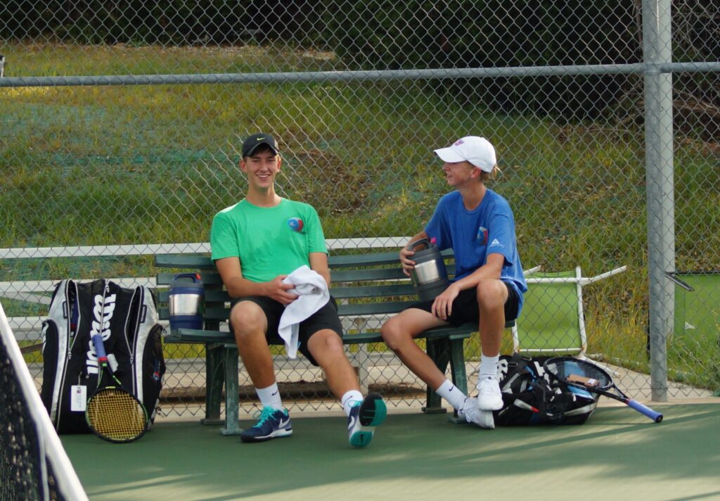 Two boys in tennis gear sit and laugh on a bench by a court, holding water jugs and tennis bags nearby.