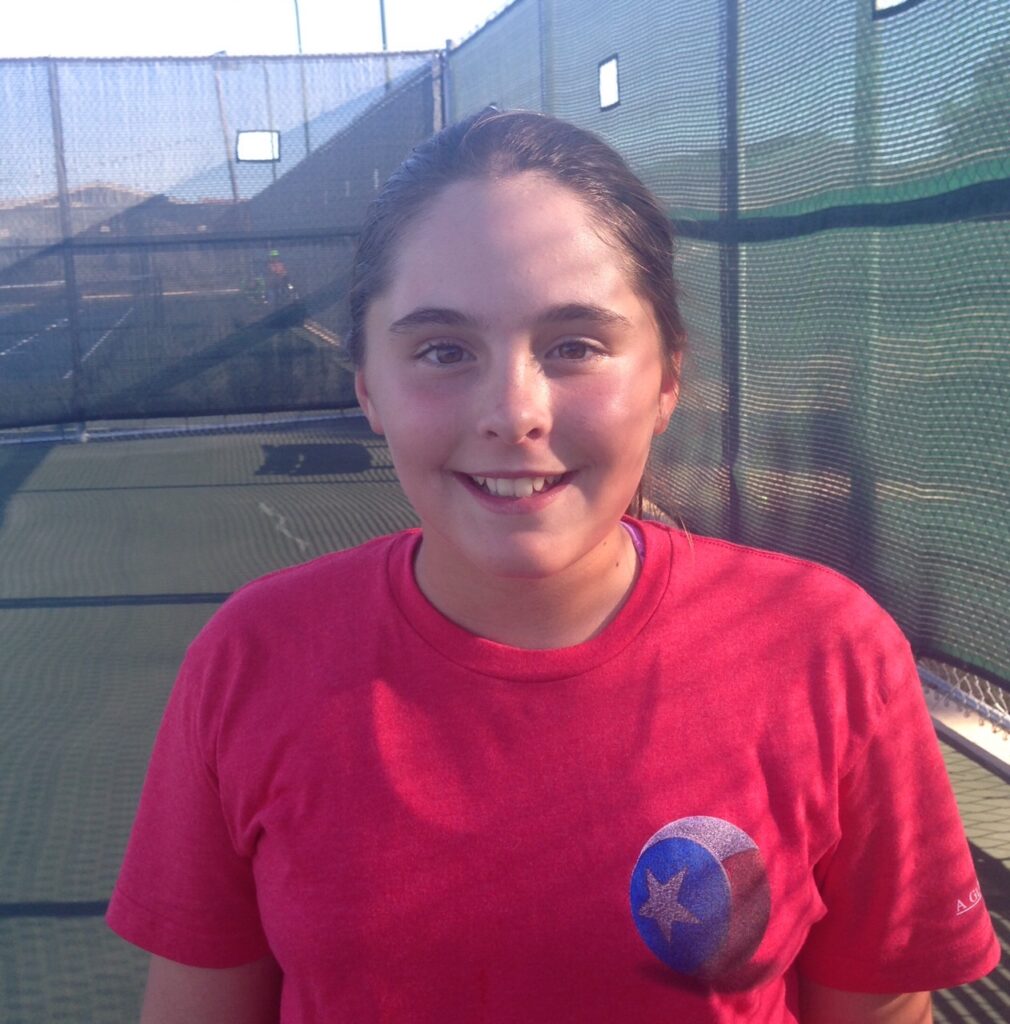 Smiling girl in a red t-shirt standing on an outdoor tennis court with green fencing in the background.