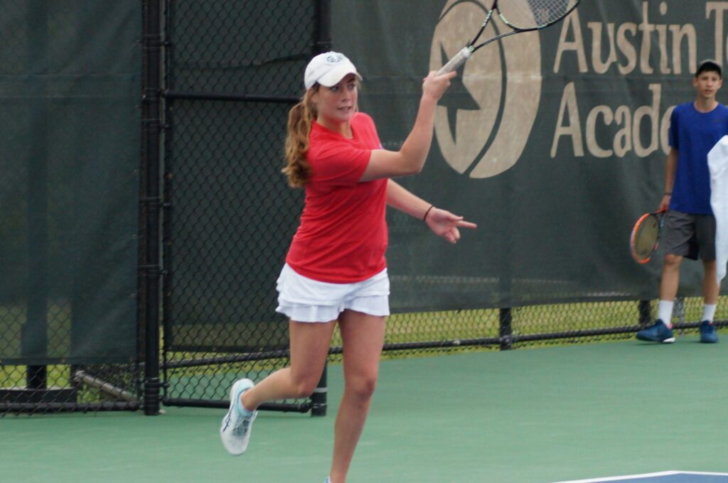 Girl in red shirt and white skirt hits a tennis ball on an outdoor court, with others visible in the background.