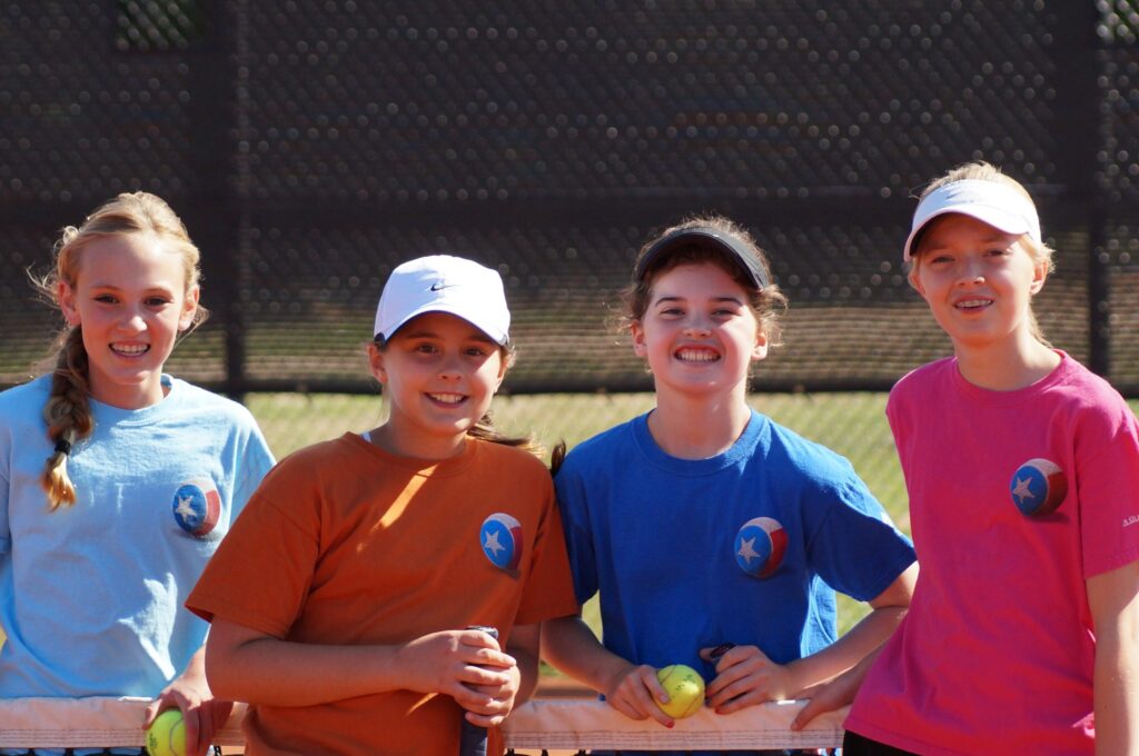 Four smiling girls in colorful shirts and hats stand together on a tennis court, holding tennis balls.