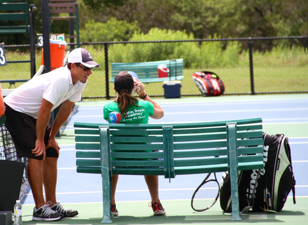 Tennis coach talks to a young player sitting on a green bench during a break on an outdoor court.