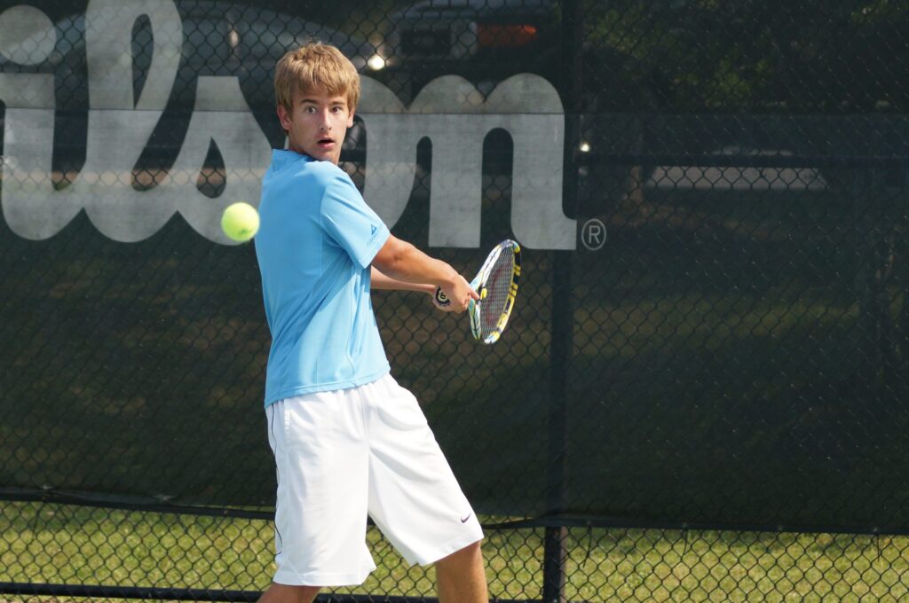 A young man in a blue shirt hits a tennis ball with a racquet on an outdoor court.
