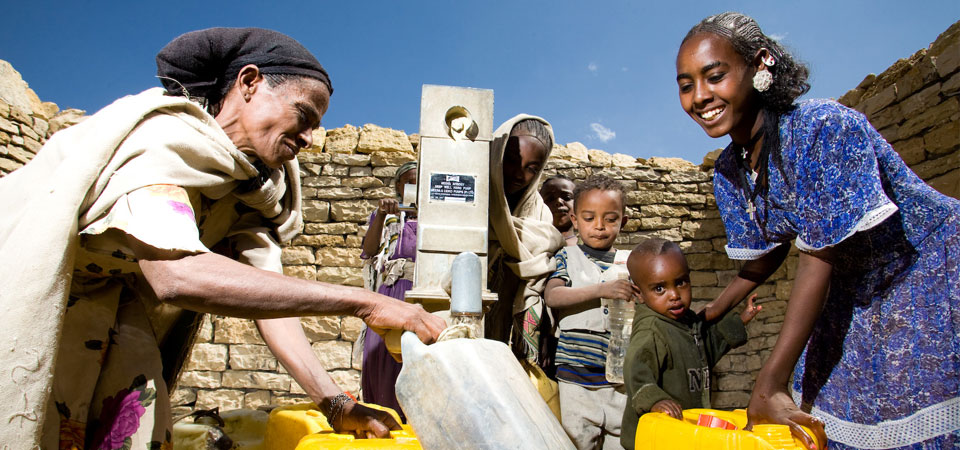 Women and children gather water from a well, filling yellow containers, smiling under a bright blue sky.