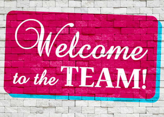 Pink sign on a white brick wall says Welcome to the TEAM! in bold white letters.