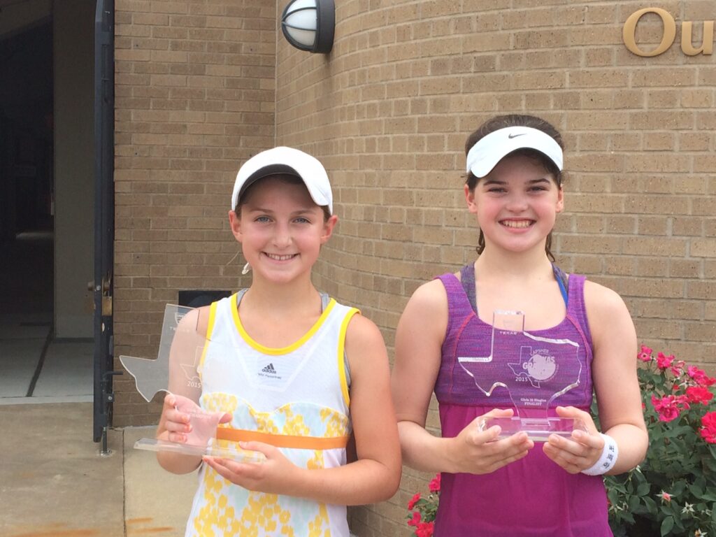 Two young girls in tennis outfits smiling and holding Texas-shaped trophies in front of a brick building.